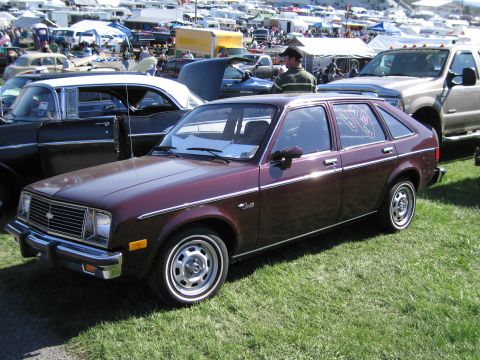 1991 Ford LTD Crown Victoria - A maroon hatchback with chrome trim and a 'Chevette' badge is parked on grass at an outdoor event with many other vehicles and tents in the…