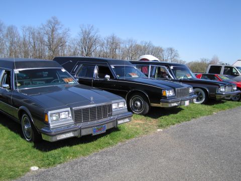 1991 Ford LTD Crown Victoria - A row of hearses and other cars are parked on grass under a clear blue sky.