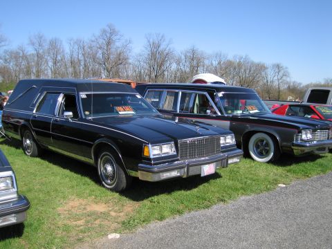 1991 Ford LTD Crown Victoria - Two vehicles, a hearse and a sedan, are parked on a grassy lot under a clear blue sky.