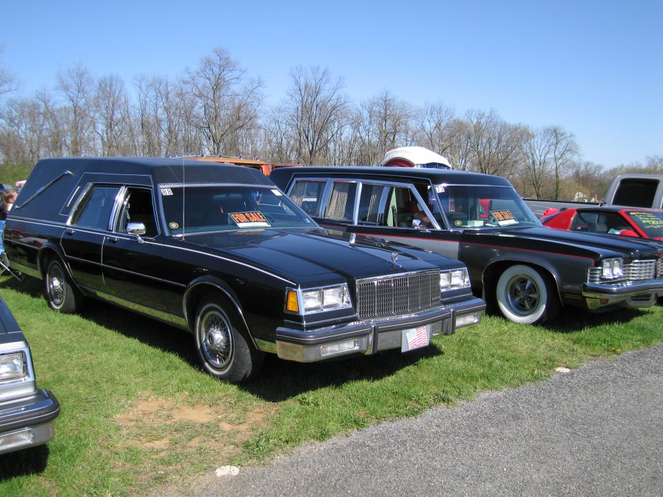 Black hearse and a dark sedan parked on grass with trees in background. 'FOR SALE' signs visible in windows