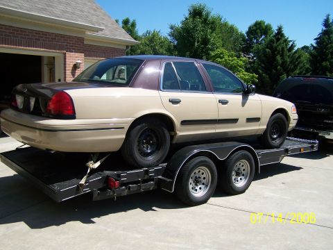 2003 Ford Crown Victoria - A 2003 Ford Crown Victoria is loaded onto a trailer, with a garage and trees in the background.