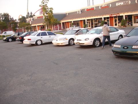 1993 Ford Taurus - Multiple Ford Taurus sedans are parked in front of a strip mall with a roller coaster in the background.
