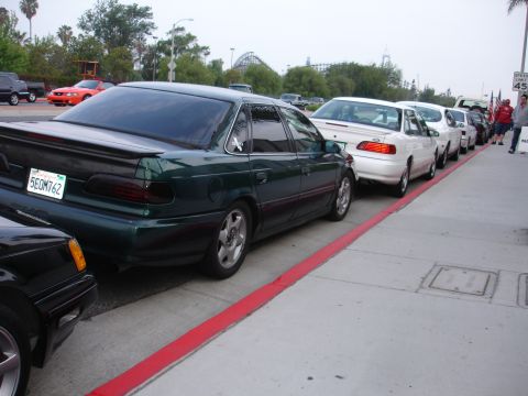 1993 Ford Taurus - A line of cars parked along a curb, with a green sedan in the foreground.