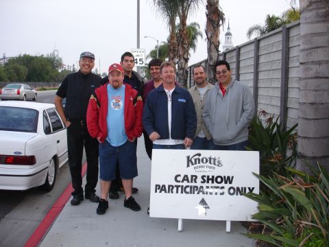 1993 Ford Taurus - A group of men stand next to a white 1993 Ford Taurus sedan at a car show.
