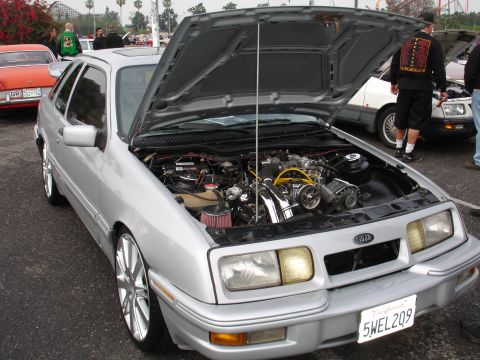 1993 Ford Taurus - The front of a silver Ford Sierra XR4Ti is shown with its hood open, revealing the engine bay. The car has custom wheels and a California l…