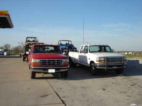 1992 Ford Bronco - A red Ford pickup truck from the early 1990s is seen from the front, towing a jeep on a trailer.