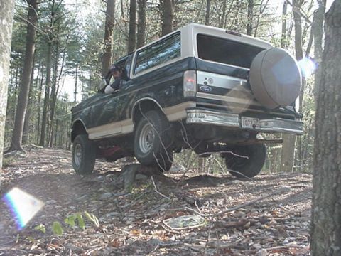 1993 Ford Bronco - 1993 Ford Bronco navigates a wooded trail, showing its off-road capabilities with a spare tire mounted at the rear.