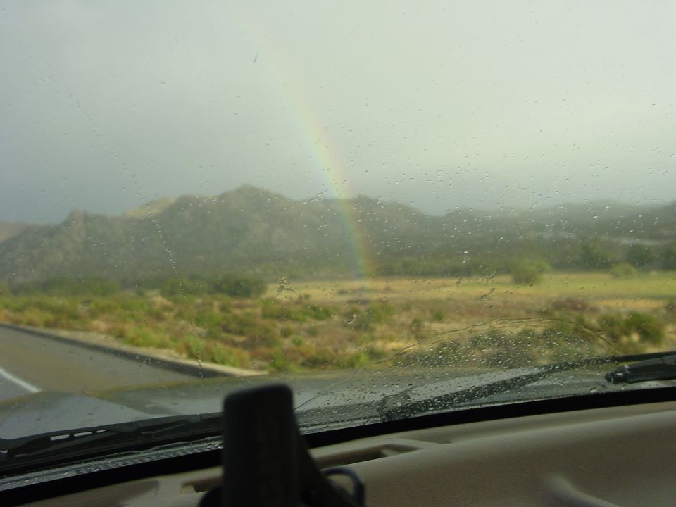 View of mountains and a rainbow through a windshield covered in raindrops. Supermotors.net logo visible