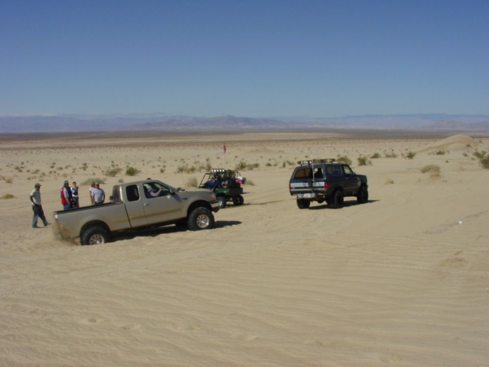 Pickup truck stuck in sand with people around it; SUV and ATV in background