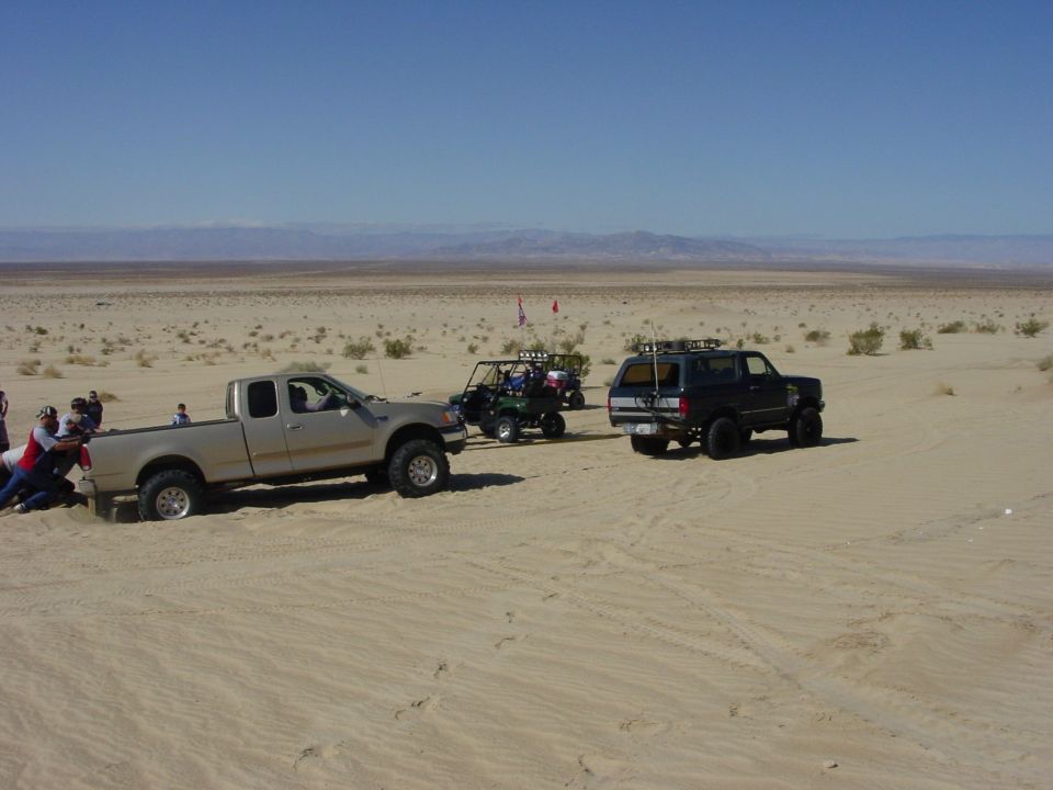 Pickup truck stuck in sand, people pushing it, another truck and a buggy behind it in desert