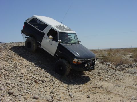 1994 Ford Bronco - Dark blue Ford Bronco with a white hardtop and white door descending a gravel berm in open desert, driver visible through the door opening…