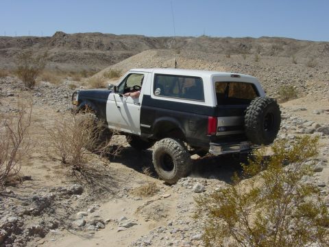 1994 Ford Bronco - Driver-side 3/4-rear view of a dark blue Ford Bronco with a white hardtop and white door, articulating through a rocky desert wash with the…