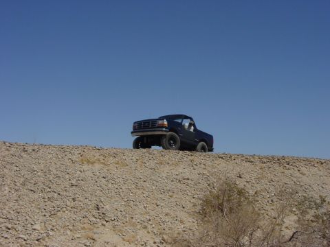 1994 Ford Bronco - A black 1994 Ford Bronco is captured from a 3/4 front view, parked on a barren, rocky landscape with a bright blue sky above.