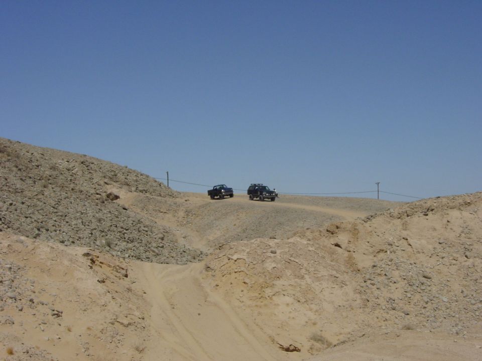 Two dark SUVs drive on a dusty dirt road through a barren, rocky landscape under a clear blue sky