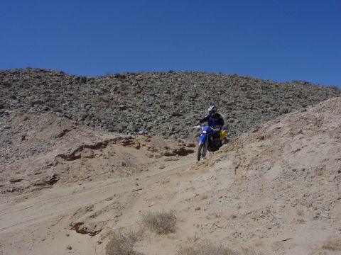 1994 Ford Bronco - A rider navigates a blue dirt bike down a sandy incline, with a rocky hillside providing a stark backdrop.