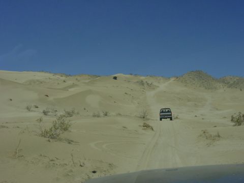 1994 Ford Bronco - A dark SUV navigates a sandy track through rolling desert dunes under a clear blue sky.