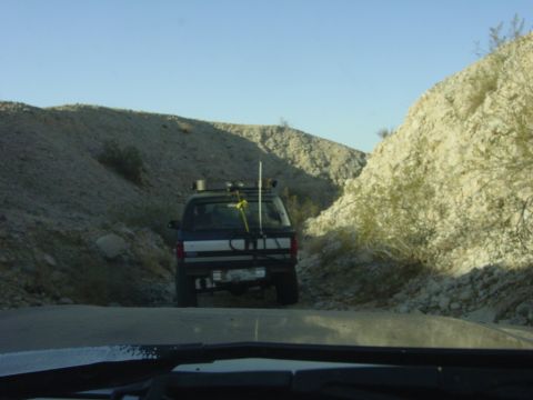 1994 Ford Bronco - An SUV travels down a dusty, narrow path through a rocky, arid landscape under a clear blue sky.