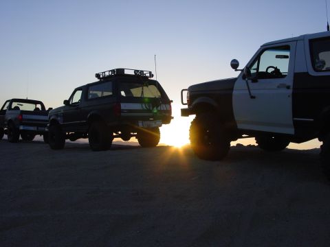 1994 Ford Bronco - Three off-road vehicles are silhouetted against the setting sun on a sandy terrain.