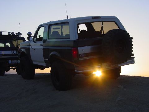 1994 Ford Bronco - A pair of modified SUVs are parked on a sandy incline as the sun sets behind them.
