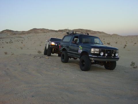1994 Ford Bronco - Two modified SUVs, a Ford Bronco and a pickup truck, traverse sandy terrain under a clear sky.