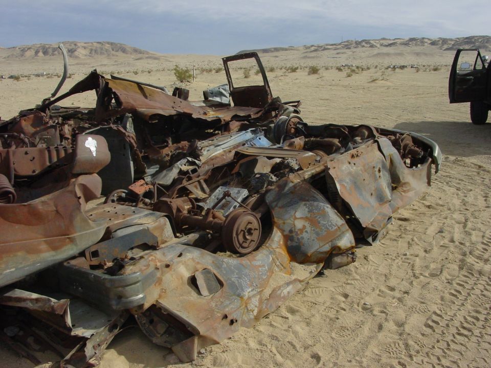 Burnt out vehicle wreckage in a desert landscape with another vehicle in the background