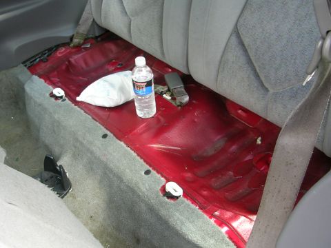 1996 Ford Probe - The interior rear floor of a vehicle shows exposed red metal and gray carpet, with a water bottle and bag on the floor.
