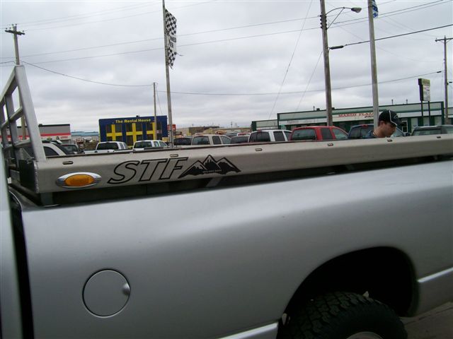 Silver pickup truck bed with STF logo and rack. Multiple vehicles in background