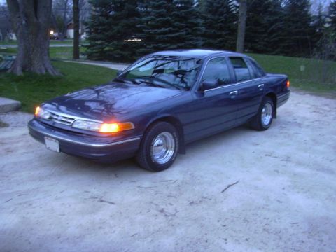 1996 Ford Crown Victoria - A dark blue sedan with chrome wheels is parked on a gravel surface, with trees in the background.