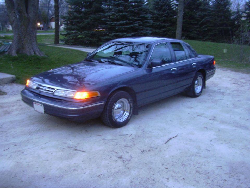 Dark blue sedan parked on a gravel driveway, front 3/4 view. Chrome wheels and amber turn signals are visible