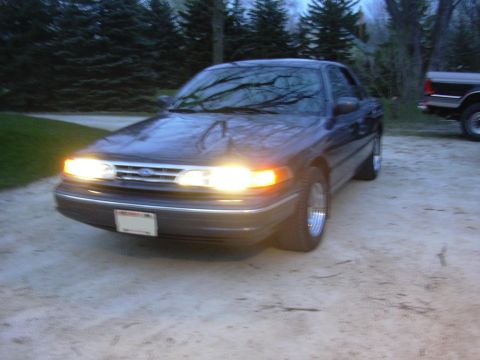 1996 Ford Crown Victoria - A dark blue sedan is parked outside with its headlights illuminated, with another vehicle partially visible in the background.