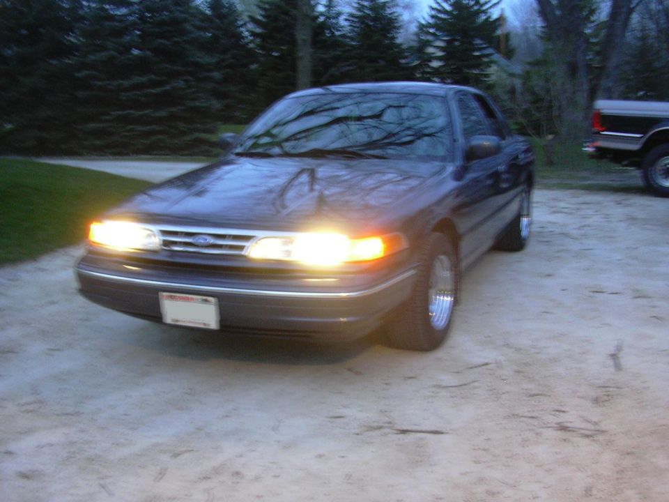Dark blue Ford Crown Victoria with headlights on, parked on a gravel driveway. Another vehicle is visible to the right