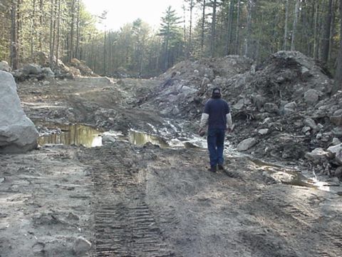 1993 Ford Bronco - person in jeans stands on a churned-up dirt track amid mud, rock piles and pine forest at an excavation site.