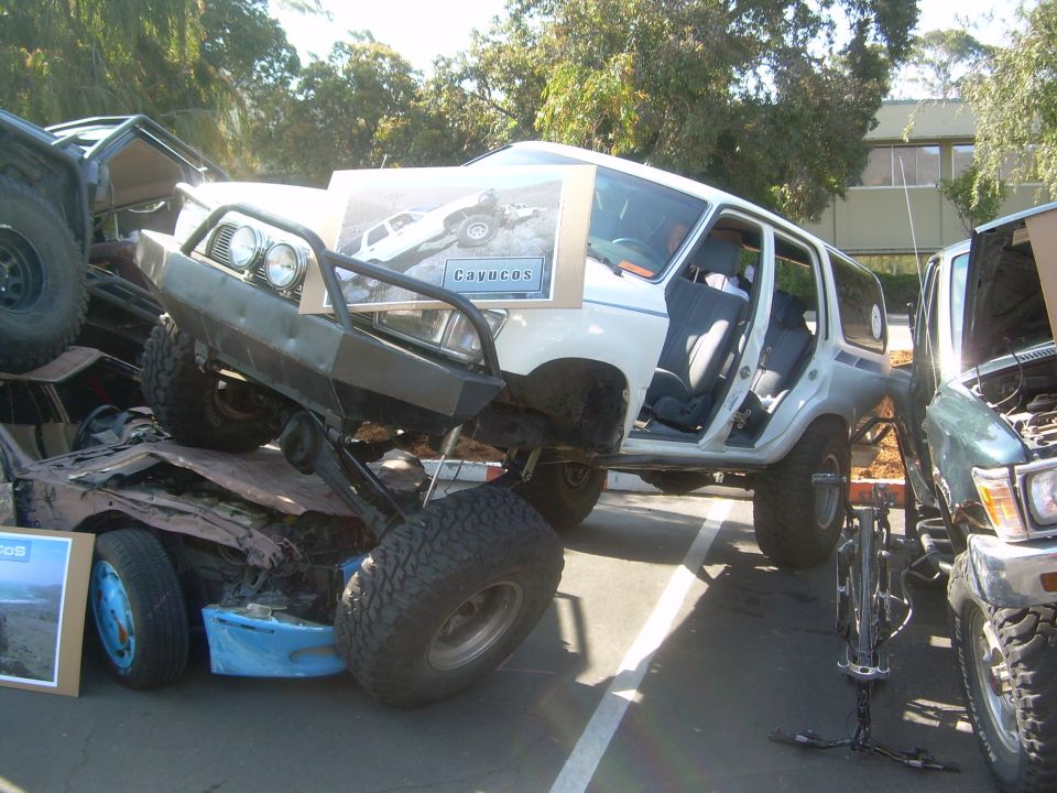 Two modified SUVs are parked close together, one on top of a crushed car. A picture is taped to the front of the top SUV