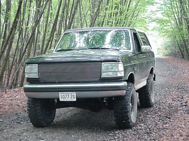 Dark green Ford Bronco parked on a dirt road with trees on both sides. License plate reads 1077 ZG
