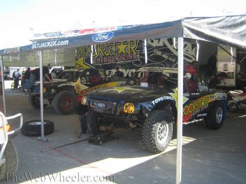 1990 Ford Bronco - Two black Rockstar Energy Drink off-road race trucks parked under a Skyjacker pit canopy with crew working nearby.