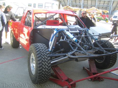 1990 Ford Bronco - red off-road race truck on a jack with its exposed rear tube frame and blue coilover shocks while crew stand nearby.