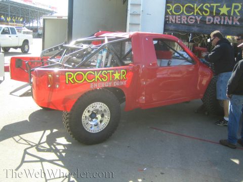 1990 Ford Bronco - red off-road race truck with Rockstar Energy Drink graphics sits in a pit area as crew members work on it.