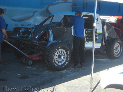 1990 Ford Bronco - Crew members in blue shirts work on a blue and white number 9 off-road race truck raised on floor jacks in a pit area.