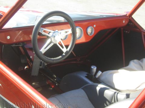1990 Ford Bronco - cockpit of a red off-road buggy showing a black three-spoke steering wheel, round gauges, shifter and bench-style seating.