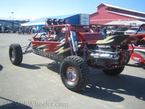 1990 Ford Bronco - maroon long-travel off-road buggy with flame graphics, roof light bar and exposed V8 engine parked on pavement at an event.