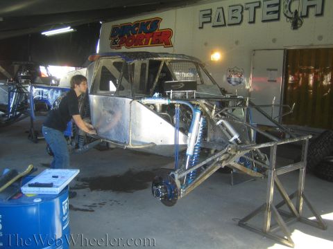 1990 Ford Bronco - man works on a bare aluminum-bodied off-road race truck with blue shocks under Dircks & Porter and Fabtech pit banners.