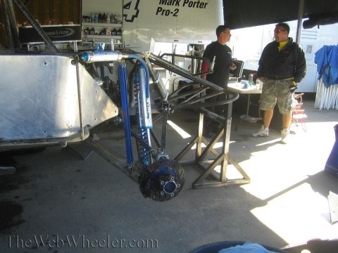 1990 Ford Bronco - Close-up of an off-road race truck's tube-frame rear suspension showing a blue coilover shock and exposed brake rotor, with men talking nea…