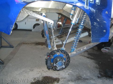 1990 Ford Bronco - Close-up of a blue off-road race truck's front suspension showing dual blue coilover shocks, A-arms and an exposed brake rotor.