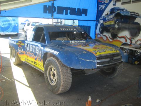 1990 Ford Bronco - mud-spattered blue off-road trophy truck wearing Toyota Tundra, Fabtech and Toyo Tires livery sits in a pit garage tent.