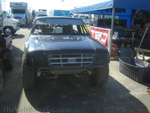 1990 Ford Bronco - flat-black off-road race truck with a Ford grille badge and tube cage sits front-on in a dusty paddock area.