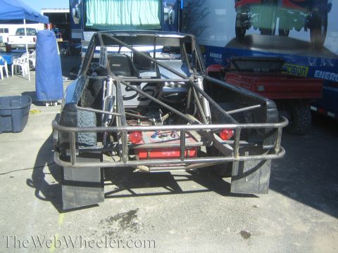 1990 Ford Bronco - bare tubular rear end of an off-road race buggy reveals the fuel cell, spare tire and rear suspension in a paddock.