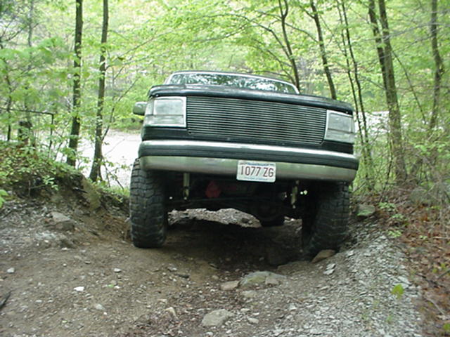 Black Ford Bronco on a dirt trail, front view. Lifted suspension, off-road tires, and a grille from the early 1990s