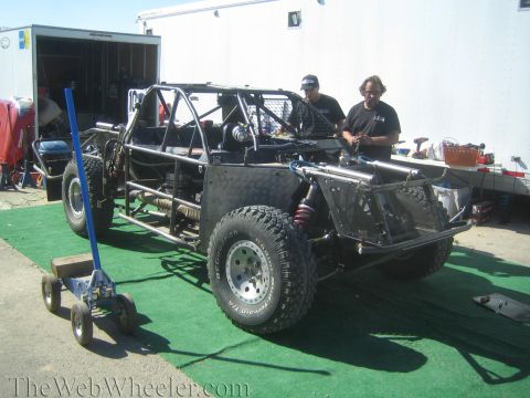 1990 Ford Bronco - Two men work on a black tube-chassis off-road race truck parked on green carpet next to an enclosed trailer.