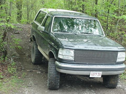 1993 Ford Bronco - Three-quarter front view of a mud-splattered dark green full-size Ford Bronco with a white hardtop flexing on an off-camber dirt trail in t…