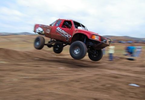 1992 Ford Explorer - A red off-road truck is airborne over a dirt ramp during a race.